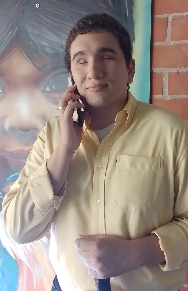 Fotografía de Santiago Luna, representante Legal de la fundación. En la imagen aparece Santiago Luna, de pie con un teléfono celular con su mano derecha y en la izquierda lleva su bastón blanco, al fondo se aprecia una cuadro o pintura. 